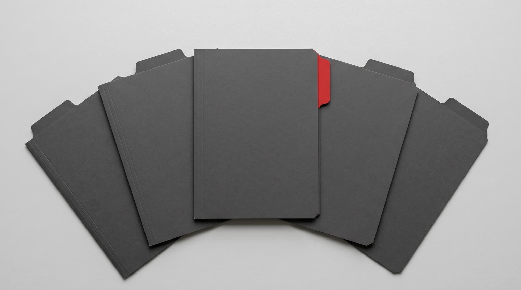 Five fanned-out dark grey tabbed index dividers on a neutral grey desk with one centre card raised slightly and marked with a single red corner tab as the only colour accent